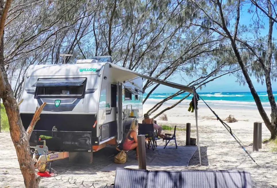 Willow RV caravan with awning out at a beachside campsite, couple relaxing in camp chairs